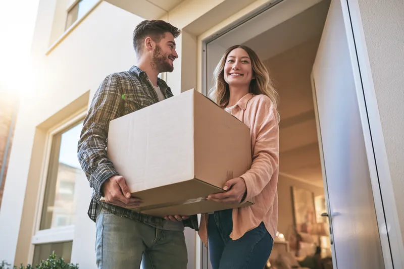 Couple in their 30s carrying a large moving box into a modern apartment building, a move often paid for with a $2,000 loan