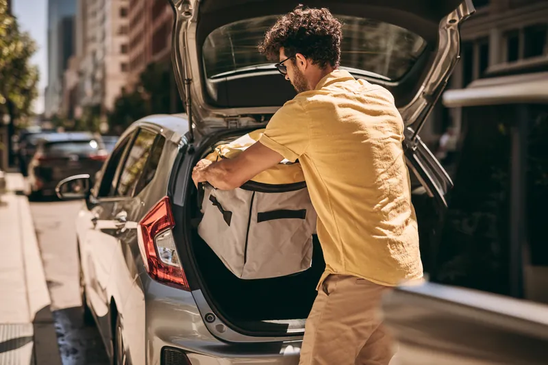 Man in his 30s lifting an insulated delivery bag from the back seat of a modern hatchback on a city street, working to grow his money stash